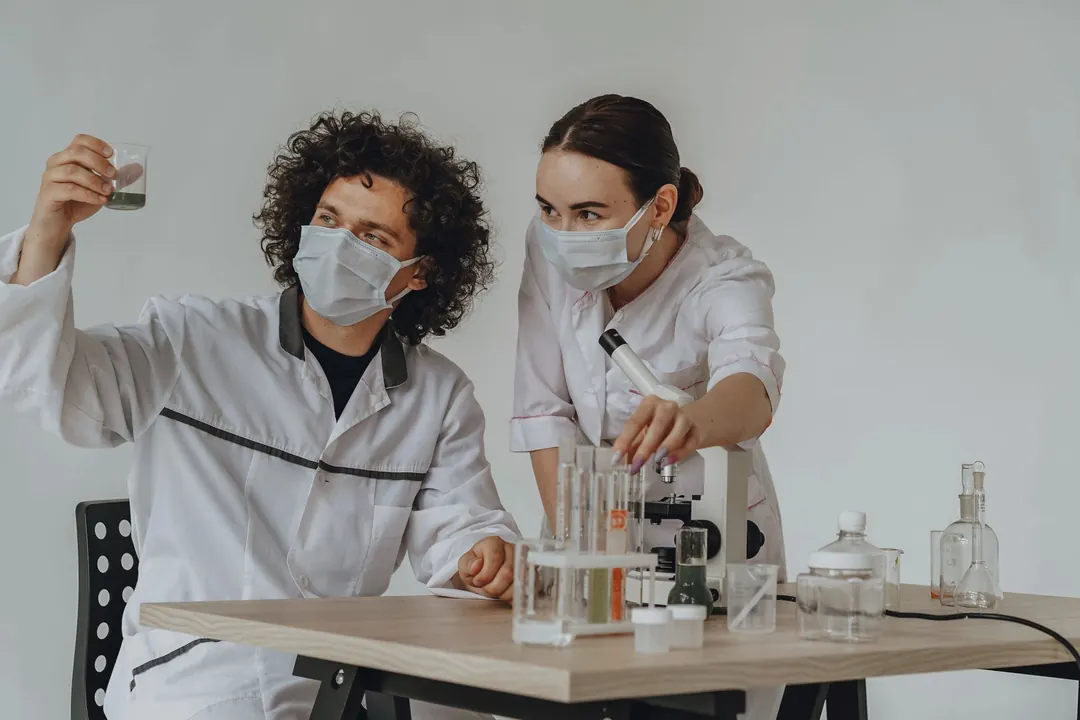 Two scientists in masks run an experiment at a bench of glassware.