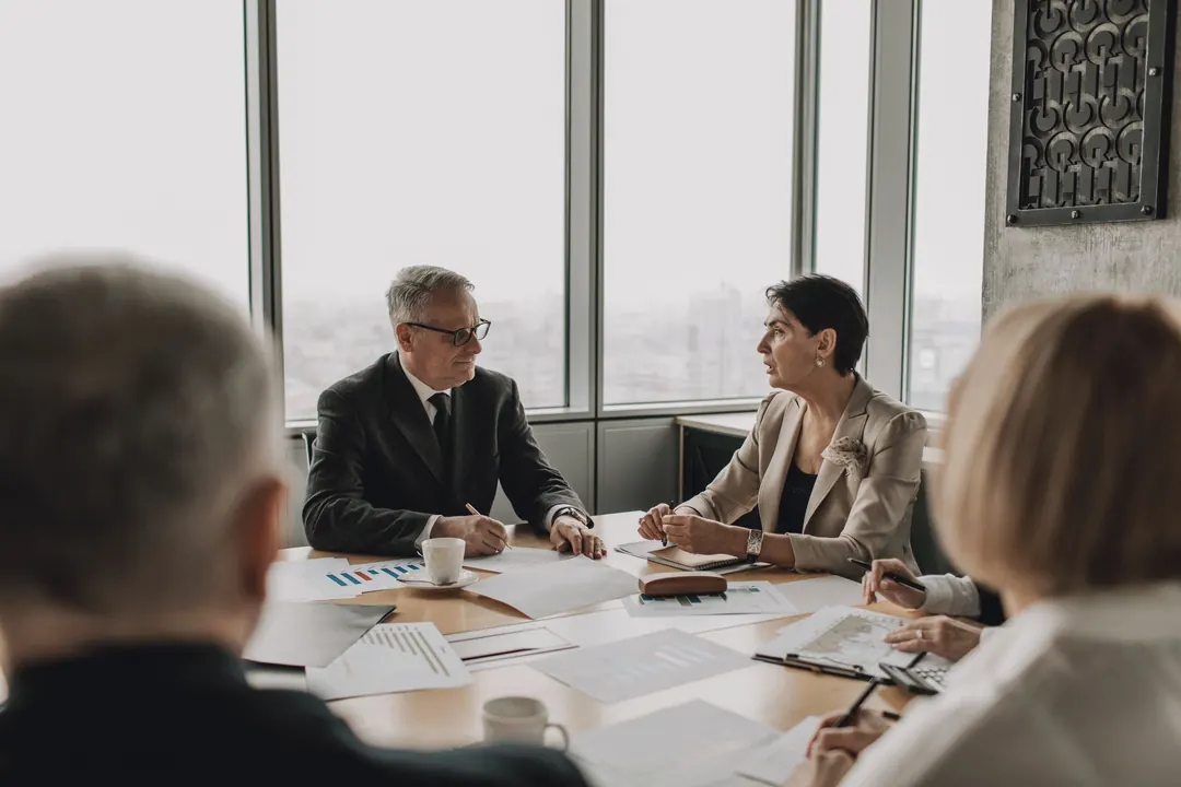 A board meets around a table by tall windows.