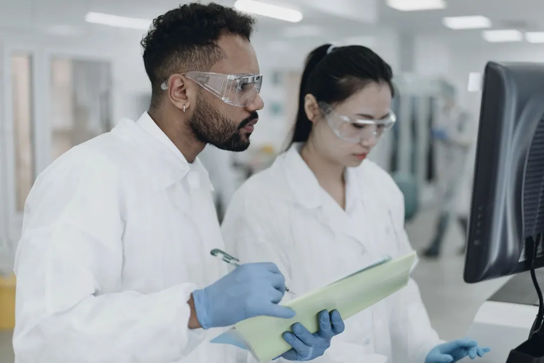 Two scientists in safety glasses check notes on a clipboard.