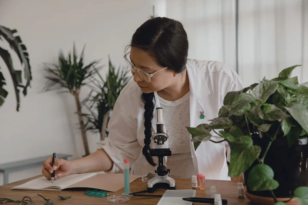 A researcher writes in a notebook beside a microscope and plants.