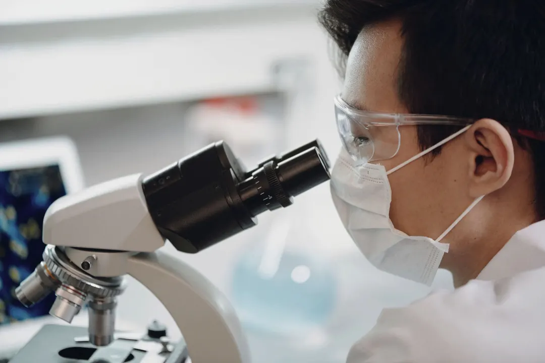 A researcher in safety glasses looks through a microscope.